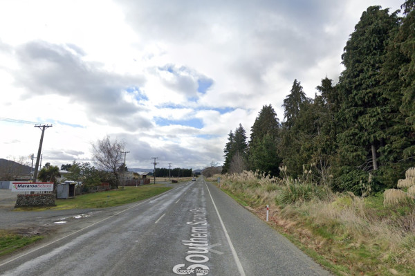 A quiet rural road with a sign for Mararoa School on the left. Tall trees line the right side under a cloudy sky, creating a serene, peaceful scene.